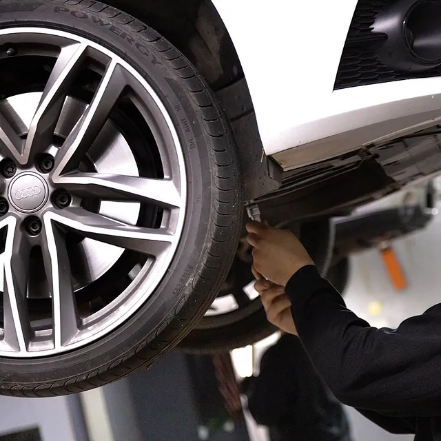 Hand eines Mechanikers, der unter einem aufgebockten weißen Auto an der Radaufhängung arbeitet