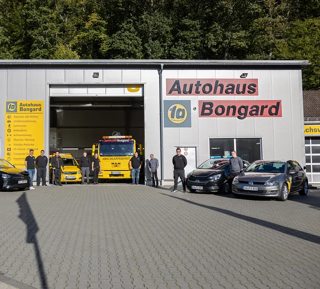 Fünf Personen stehen vor einem Autohaus mit mehreren Fahrzeugen, darunter ein gelber Abschleppwagen, vor einer hellen Fassade mit dem Schriftzug 'Autohaus Bongard' und einem gelben Firmenschild.