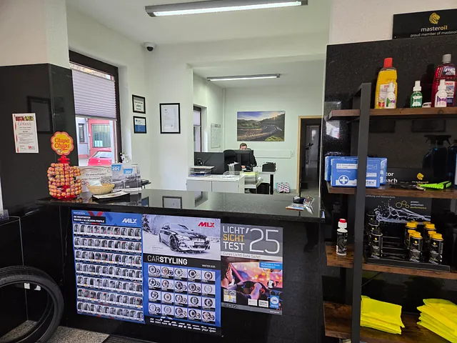Blick in eine Werkstatt mit Theke, an der Poster zu Autolichttests und Carstyling hängen, Regalen mit Autopflegeprodukten und einem Mitarbeiter am Computer im Hintergrund.