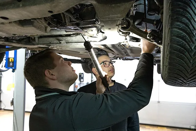 Zwei Männer inspizieren unter einem angehobenen Auto das Fahrwerk, einer hält eine Taschenlampe