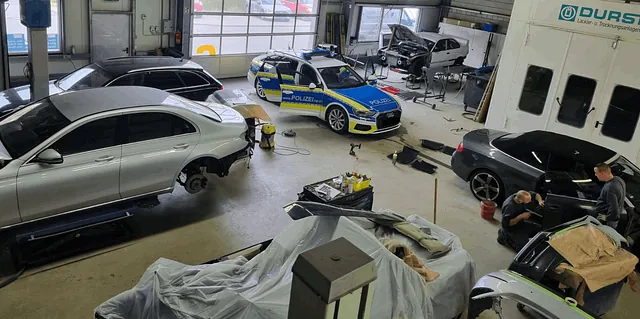 Werkstatt mit mehreren Autos, darunter ein Polizeiauto, einige Fahrzeuge ohne Räder und zwei Personen, die an einem schwarzen Auto arbeiten