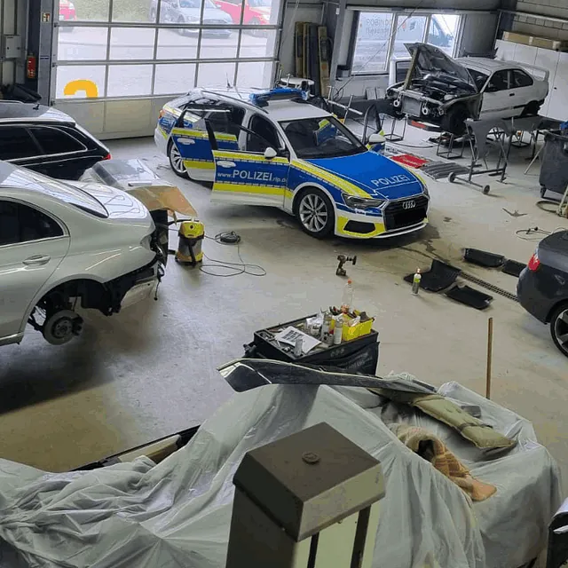 Werkstatt mit mehreren Autos, darunter ein Polizeiauto, einige Fahrzeuge ohne Räder und zwei Personen, die an einem schwarzen Auto arbeiten